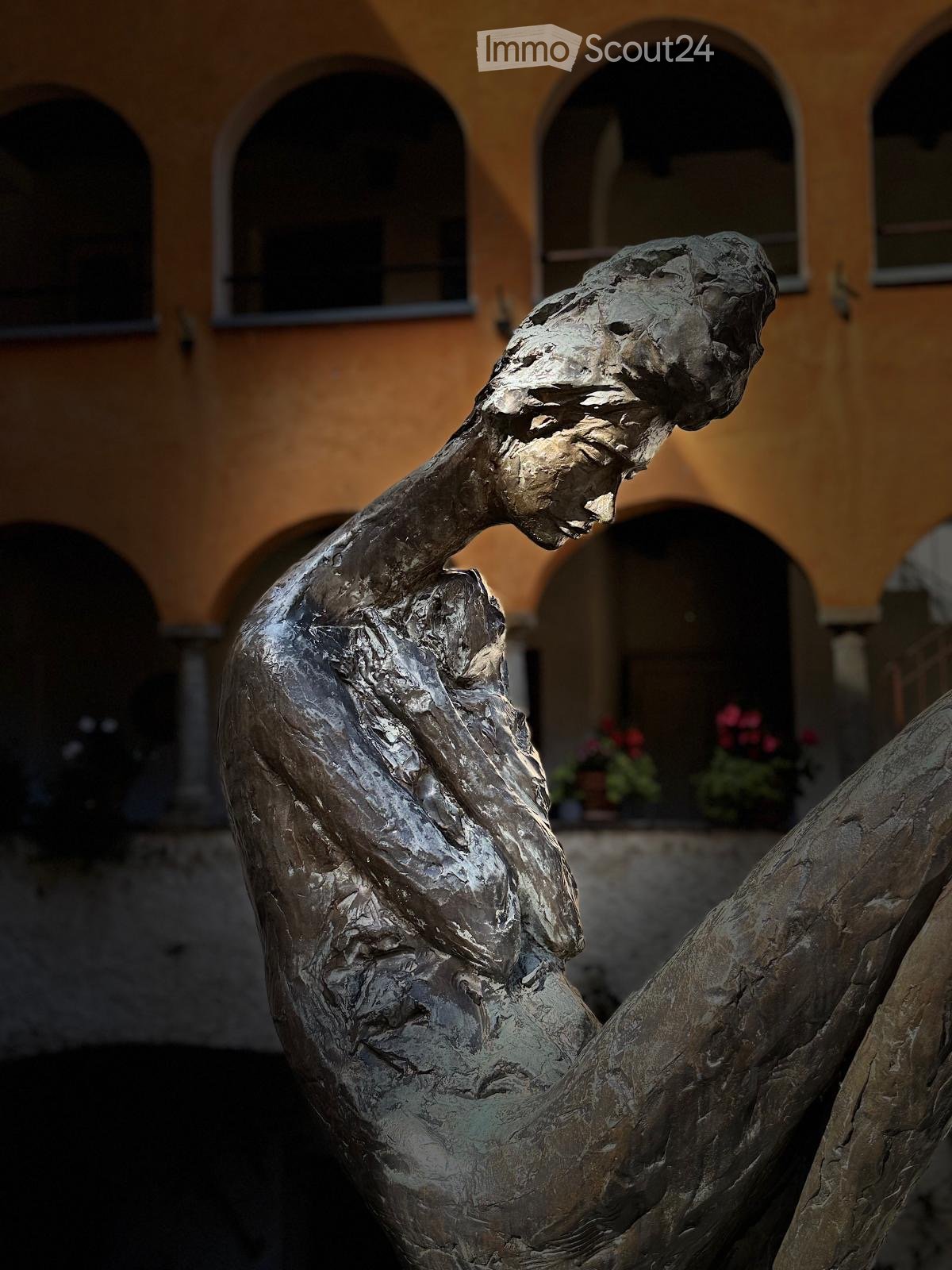 Bronze sculpture of a human figure in a dynamic pose, set against a backdrop of a historic building with arched windows and potted plants.