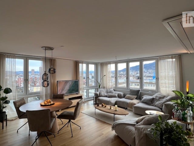 Modern living room with large windows offering a city view, featuring a grey sectional sofa, coffee table, dining table with chairs, and a television. The room has wooden flooring and pendant lights.