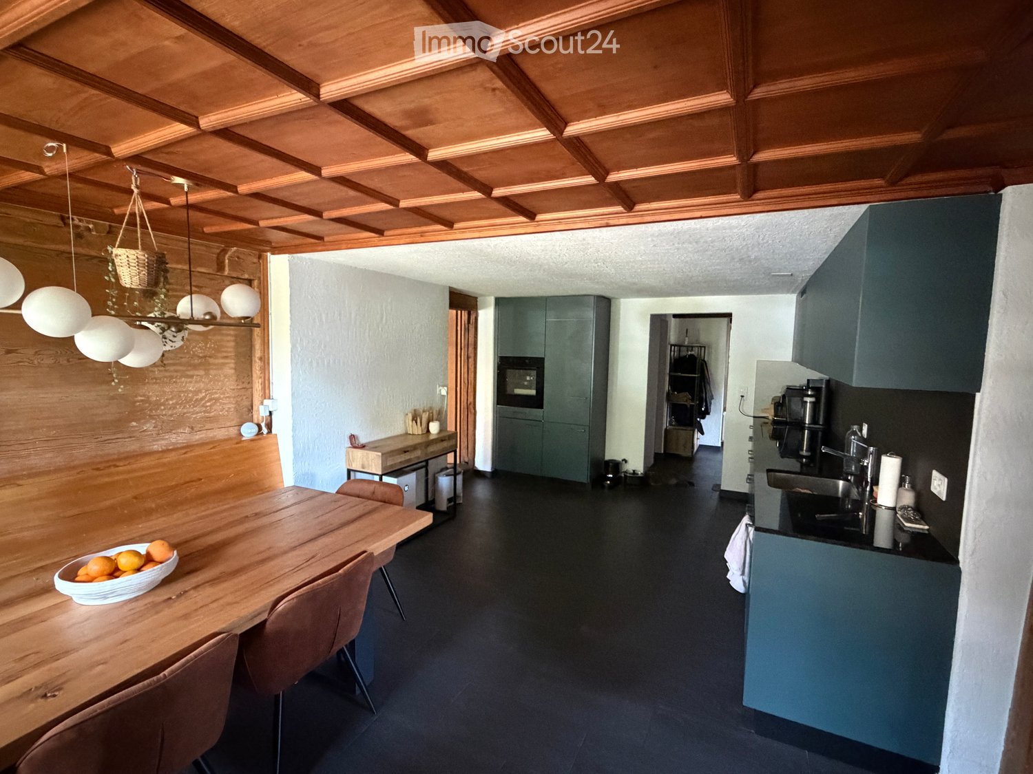 Modern kitchen with wooden ceiling, dark flooring, and grey cabinets. Features a wooden dining table with chairs, a bowl of oranges, and a hanging light fixture.