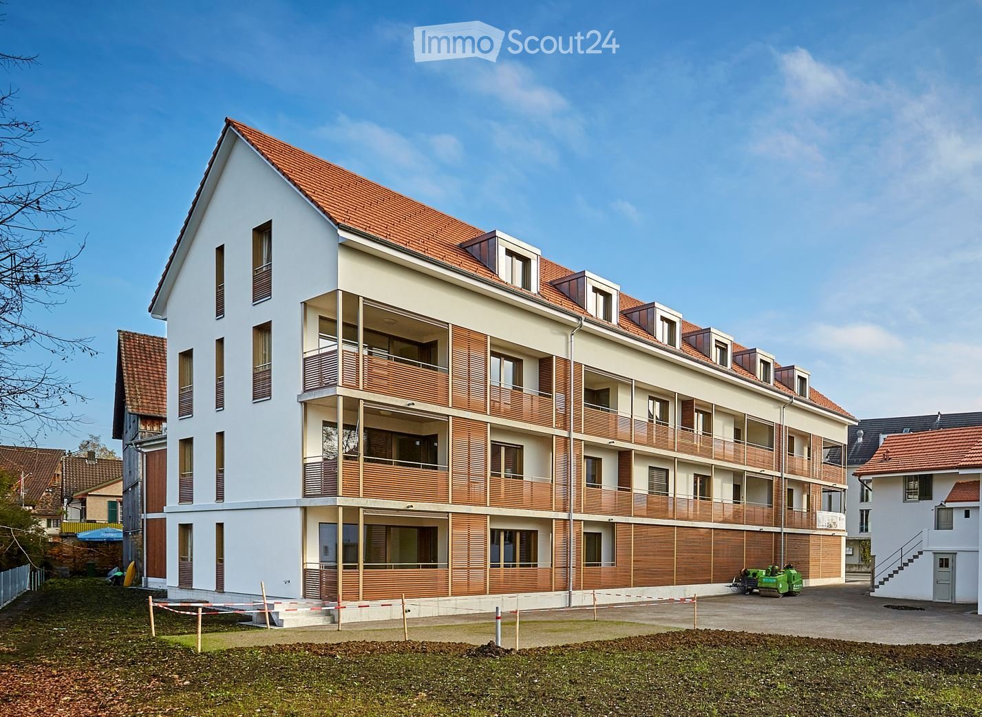 Modern multi-story residential building with white walls, wooden balconies, and red-tiled roof. The building features multiple windows and a raised ground floor entrance.