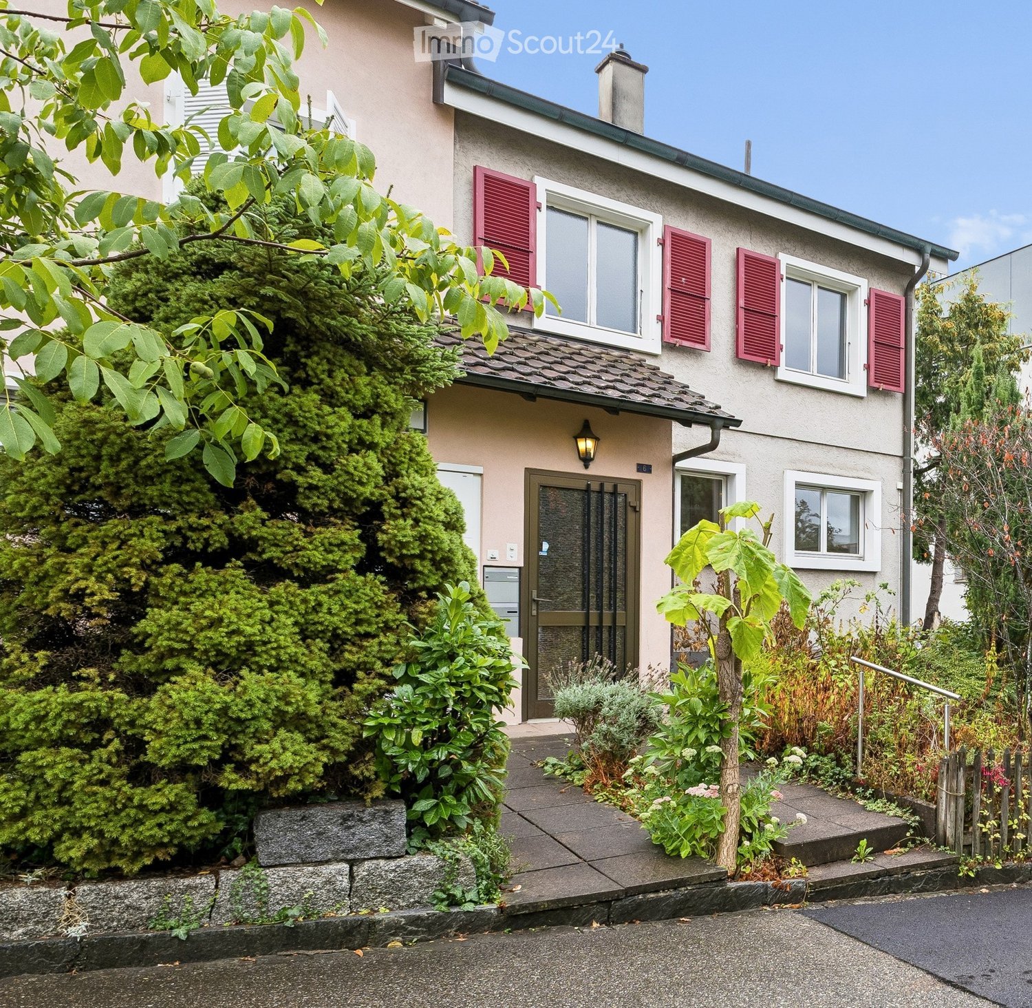 Two-story house with beige siding, red shutters, and a well-maintained garden with various plants and shrubs.
