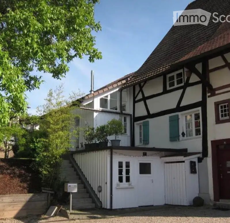 Two-story house with a raised ground floor, white siding, dark wooden beams, and green shutters. A small staircase leads up to the entrance.