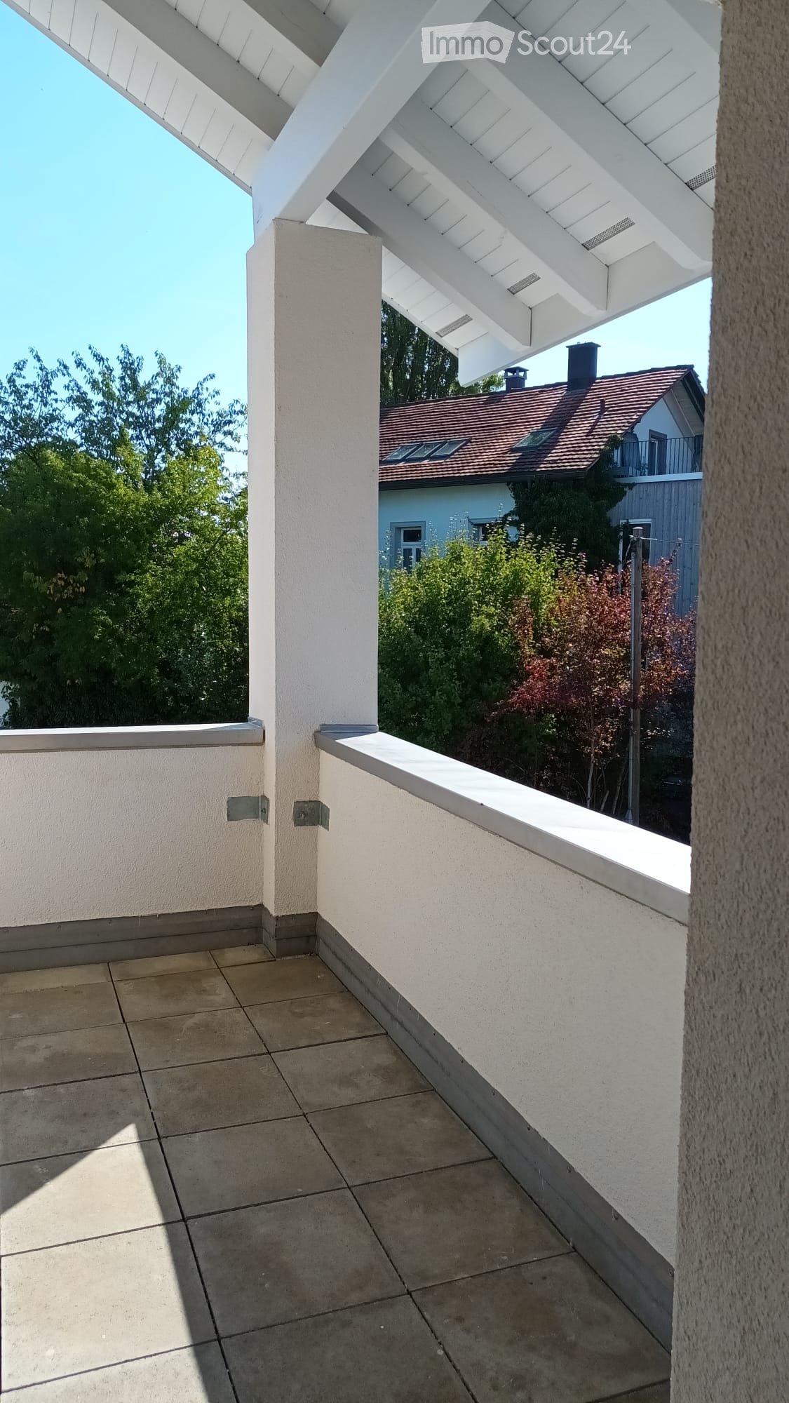 Balcony with tiled floor, white walls, and a white roof structure. The view includes a neighboring house with a red-tiled roof and surrounding greenery.