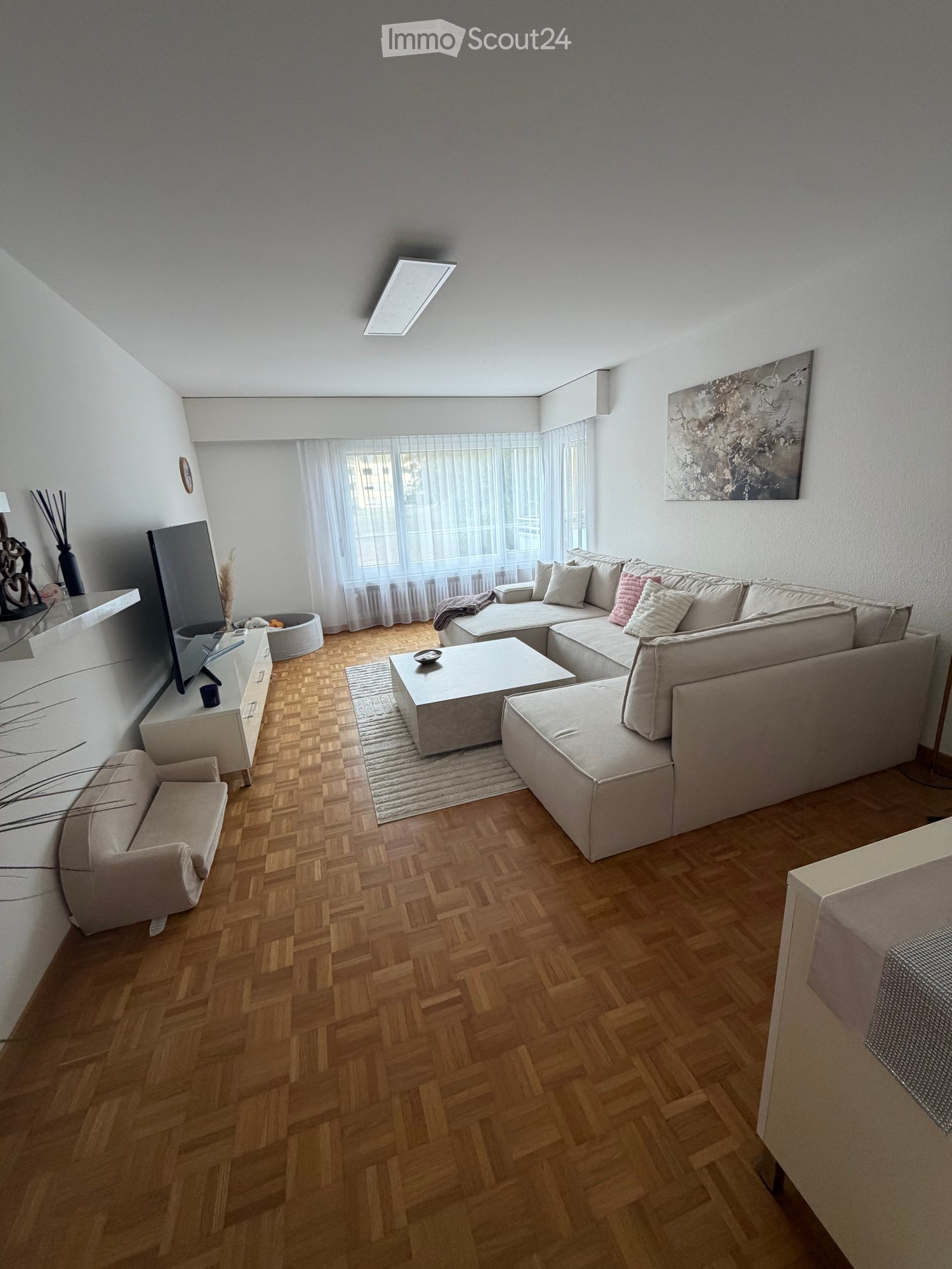 Modern living room with light-colored wooden flooring, a large sectional sofa, a coffee table, an armchair, a TV stand, and decorative items including a vase and a picture frame.