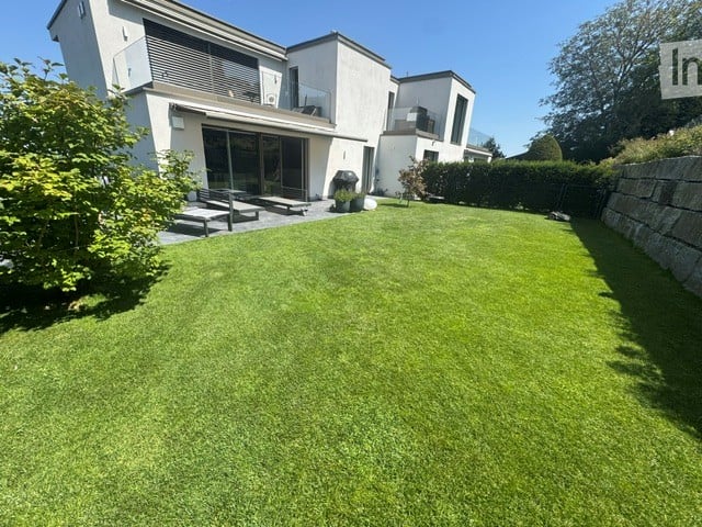 Well-maintained lawn in front of a modern house with large windows and a balcony, surrounded by greenery and a stone wall.