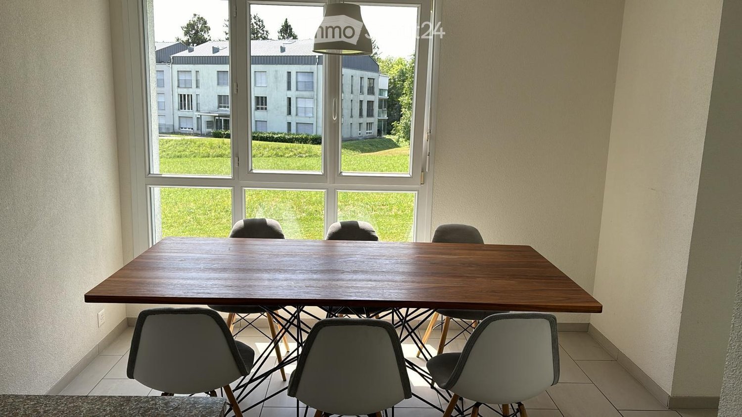 Bright room with large windows overlooking a green lawn and modern buildings. A wooden dining table is surrounded by six grey chairs.