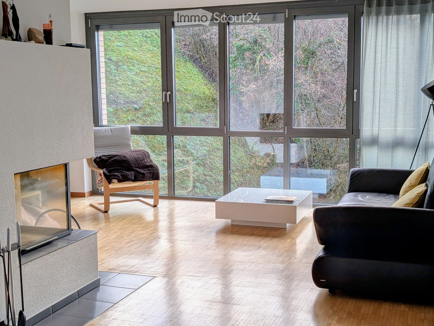 Modern living room with large windows, wooden flooring, a black leather sofa, a glass coffee table, a wooden chair with a blanket, and a fireplace.