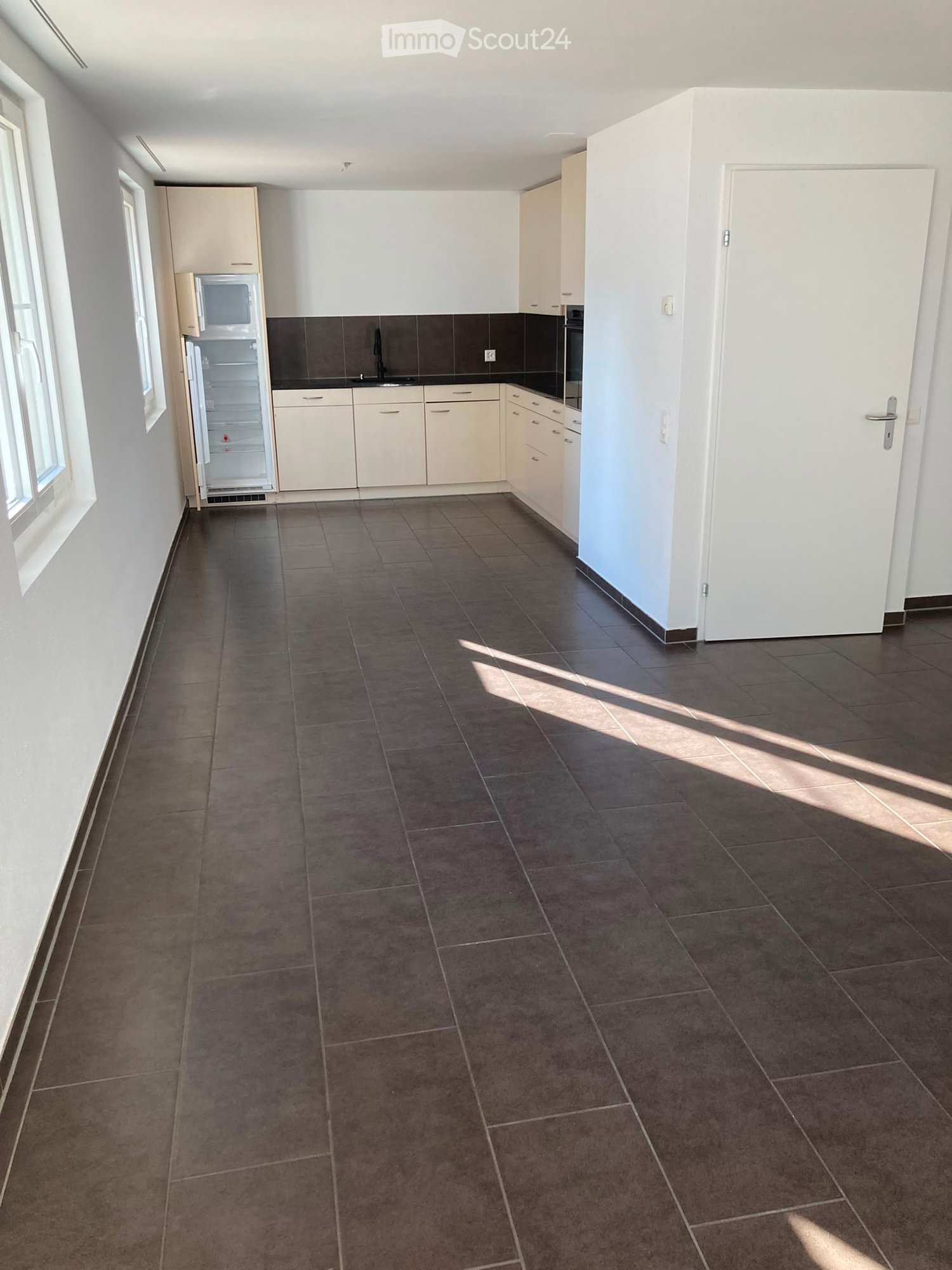 Empty kitchen with tiled floor, white cabinets, and built-in appliances including a refrigerator, microwave, and oven. Sunlight streams in from the left side.