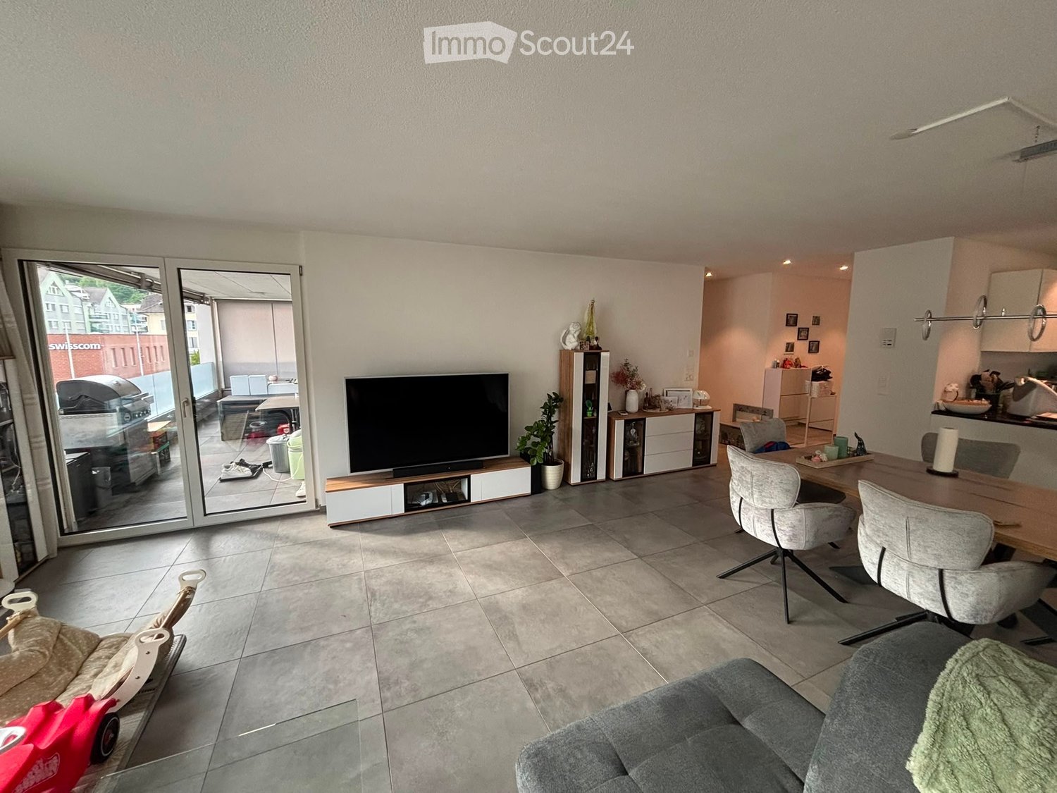 Modern living room with a grey sofa, glass coffee table, dining table with chairs, TV, cabinet, and potted plant. Large sliding glass doors lead to a balcony.