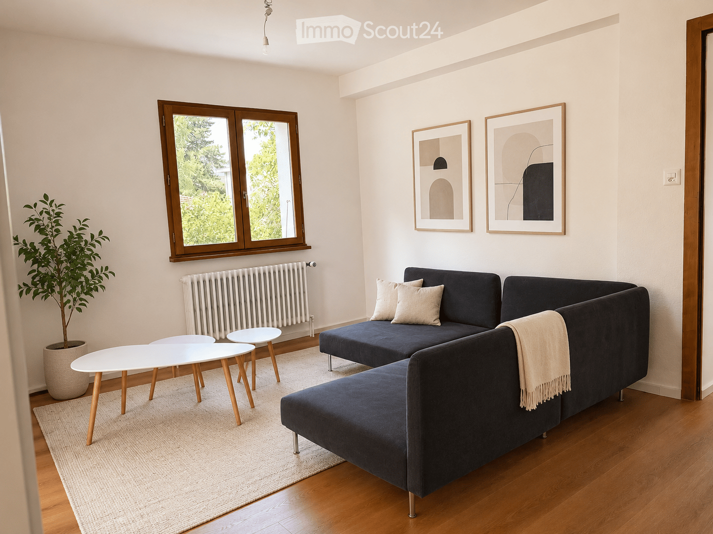 Modern living room with a dark blue sectional sofa, white coffee table, small white stools, a large plant in a pot, and two framed pictures on the wall. The room features wooden flooring and large windows with wooden frames.