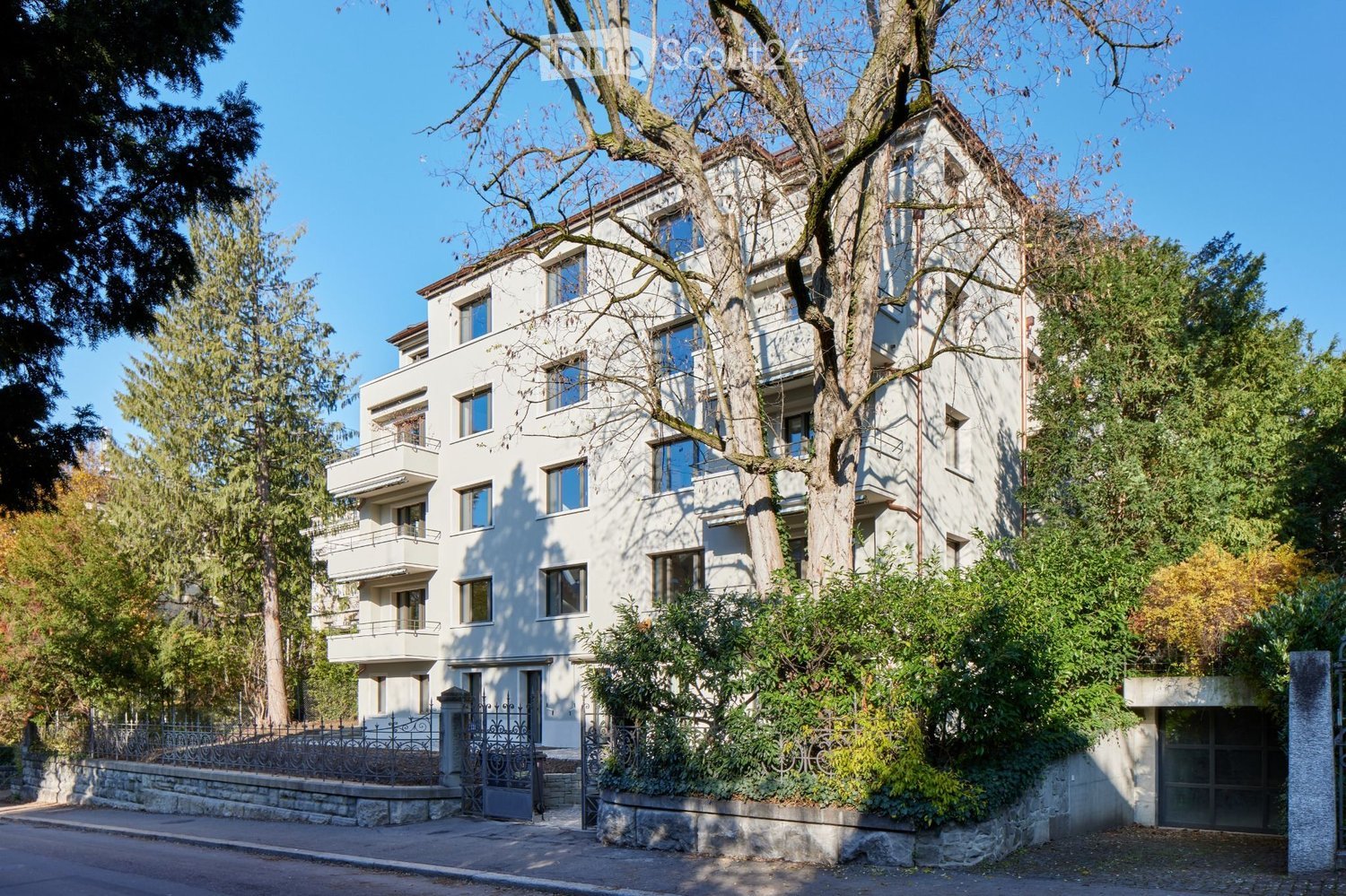 A multi-story white building with balconies, surrounded by trees and greenery, with a gated entrance and stone steps leading up to it.