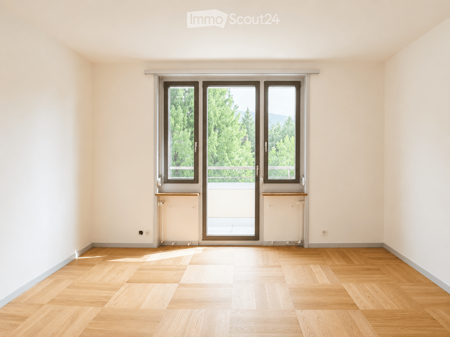 Empty room with light wooden flooring, white walls, and large sliding glass doors leading to a balcony with a view of greenery.