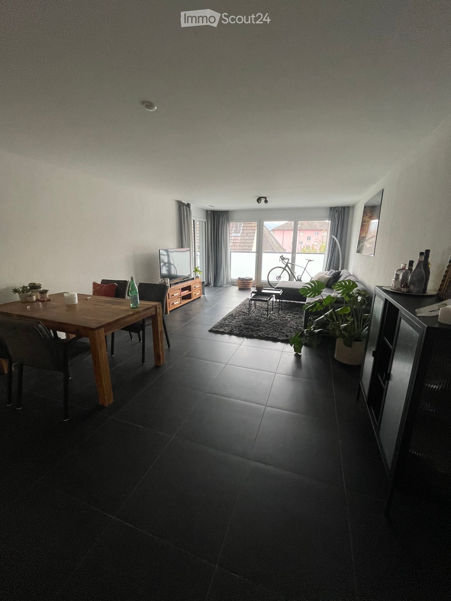 A modern, open-plan living and dining area with dark tiled flooring, a large sofa, a wooden dining table with chairs, a television on a stand, and several potted plants. The room features large windows with curtains and a view of a bicycle outside.