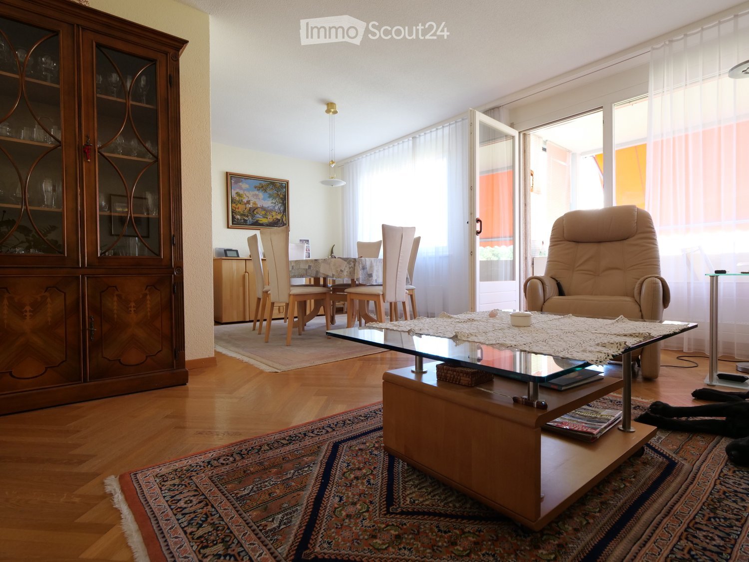 Bright living room with wooden floors, large windows, a glass coffee table, a beige sofa, a dining area with chairs and a table, and a wooden cabinet with glass doors.