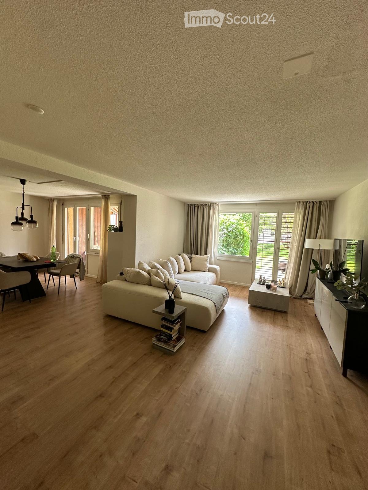Modern living room with light wooden flooring, large windows with curtains, a white sectional sofa, a coffee table, dining area with chairs and table, and decorative items on side tables.