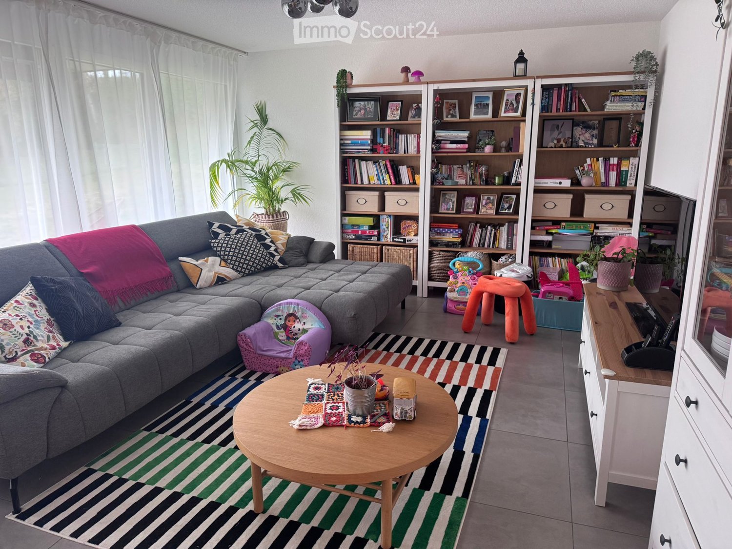 Living room with grey sectional sofa, colorful cushions, coffee table, bookshelf, and small orange stool.