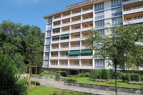 Multi-story residential building with balconies, surrounded by a landscaped garden area with trees and pathways.