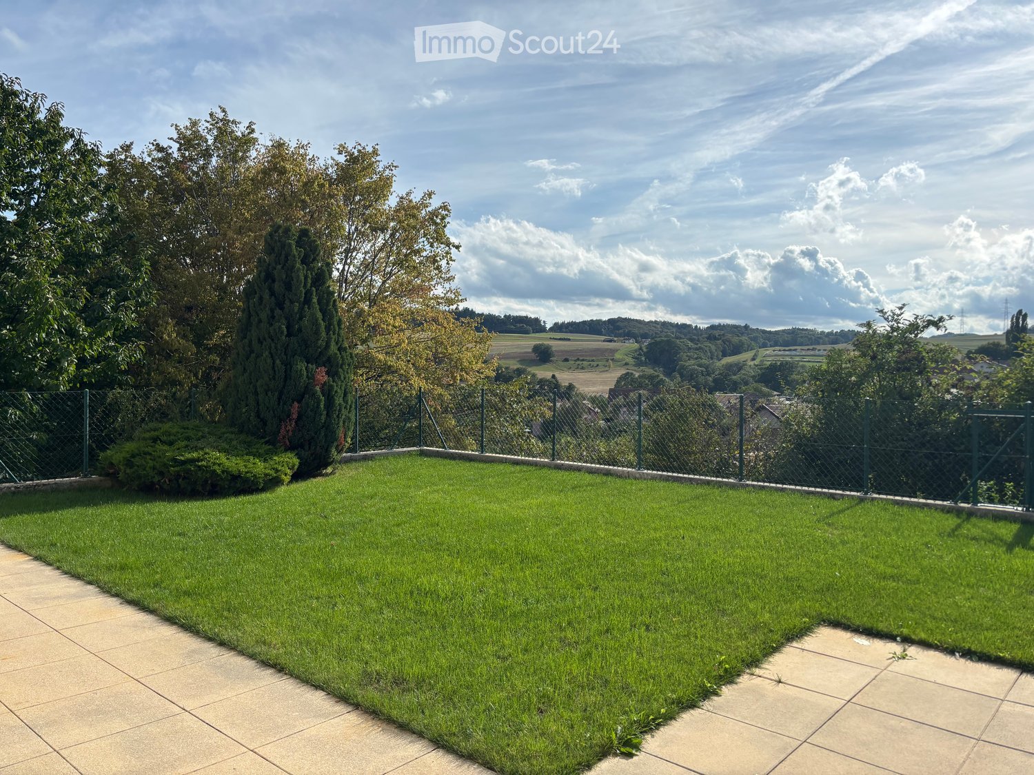 Well-maintained garden with green grass, tiled pathways, and a metal fence, offering a scenic countryside view.