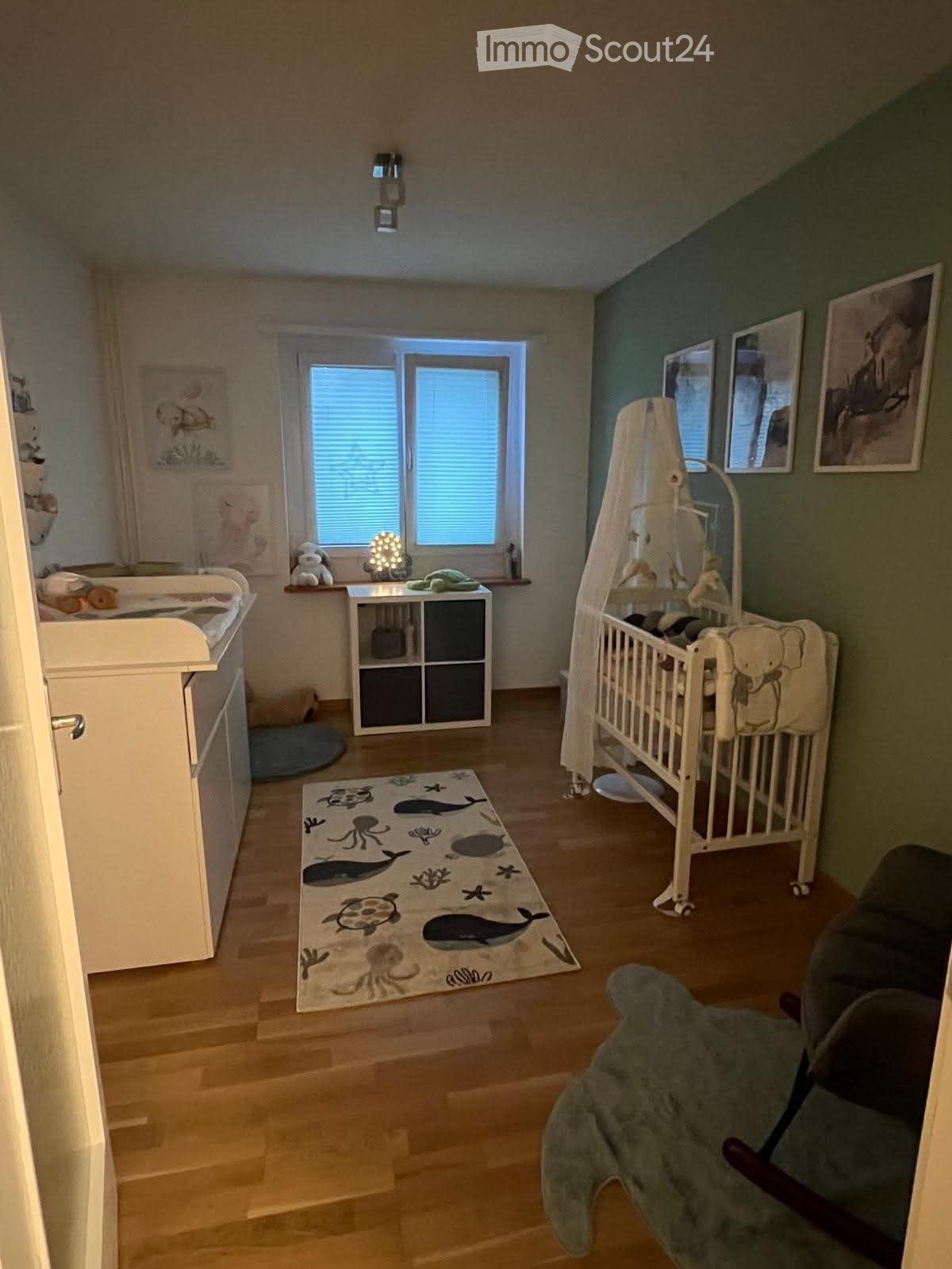 A well-lit children's bedroom with a crib, changing table, storage unit, ocean-themed rug, and picture frames on the walls.