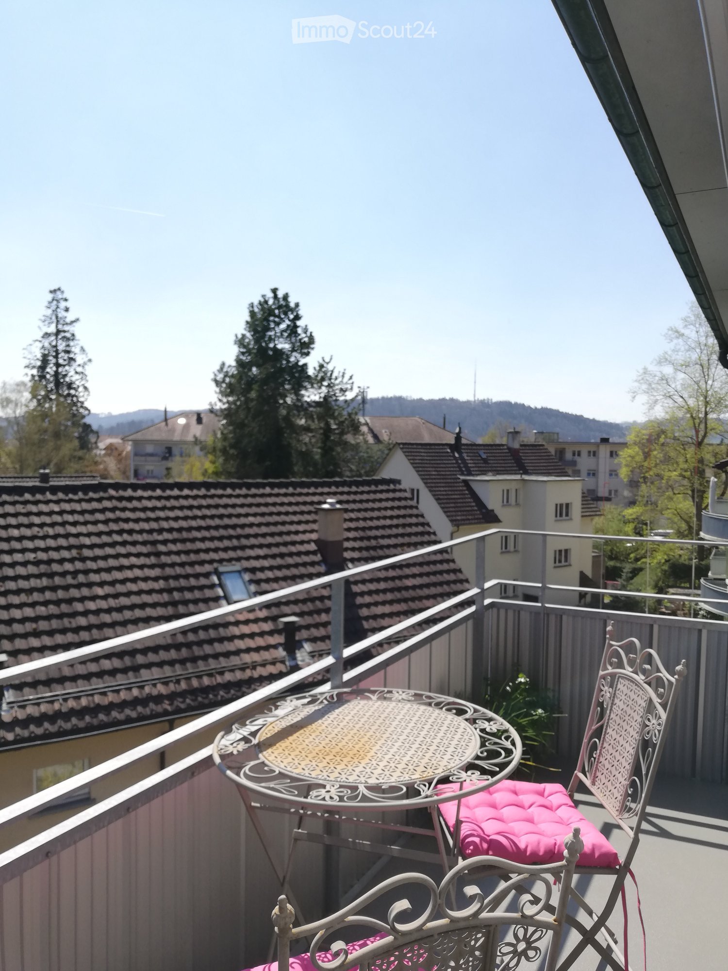 Balcony with a round metal table and two matching chairs with pink cushions, overlooking a residential area with tiled roofs and trees.