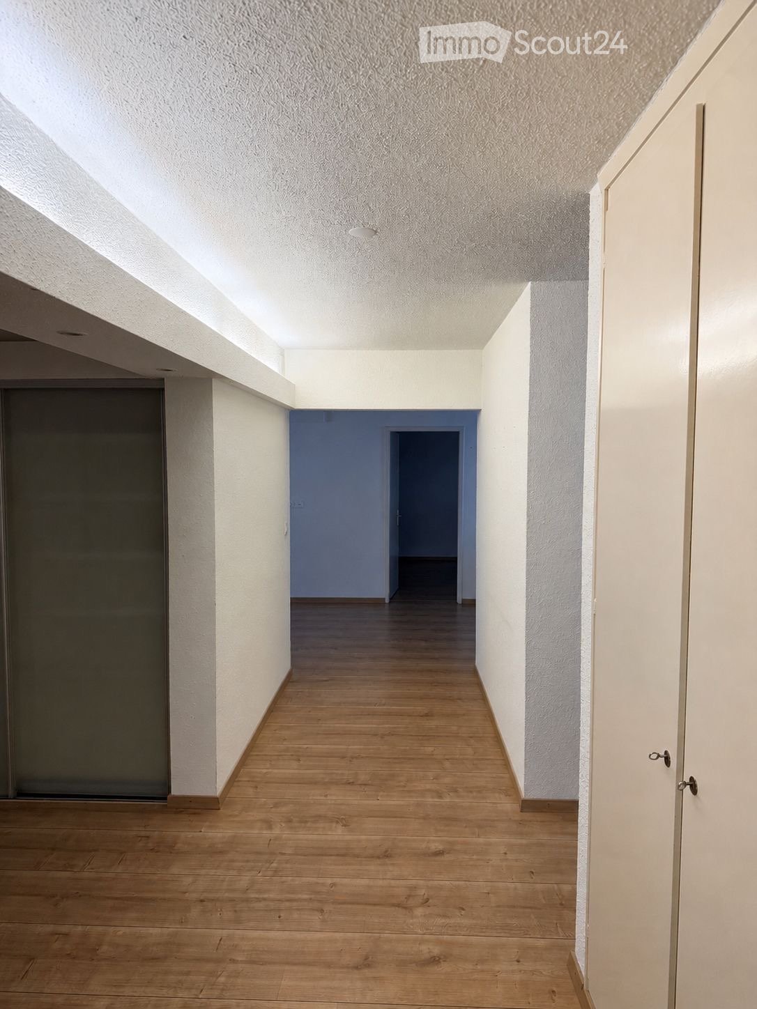 Empty hallway with wooden flooring, white walls, and a blue accent wall leading to another room.