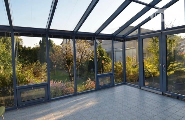 Empty sunroom with large glass walls and roof, tiled floor, and views of a garden with trees and plants.