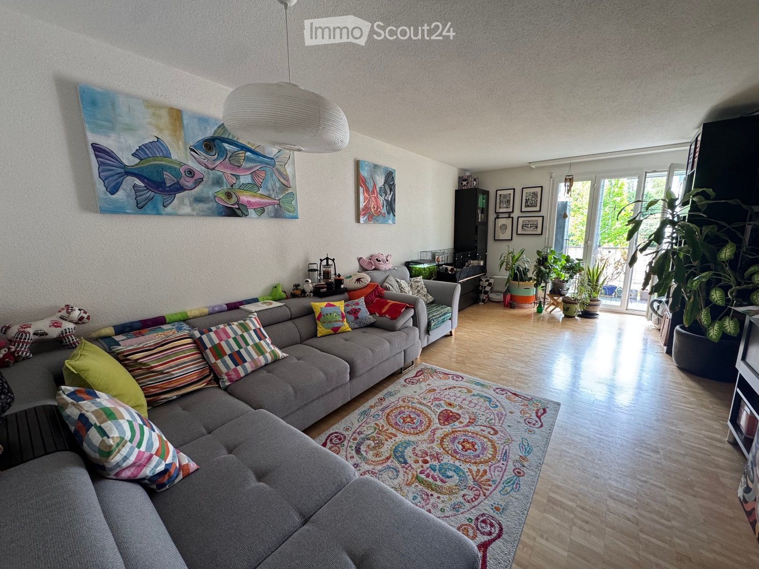 Living room with a grey sectional sofa adorned with colorful pillows, a coffee table, a large pendant light, a patterned rug, and several potted plants near the sliding glass doors.