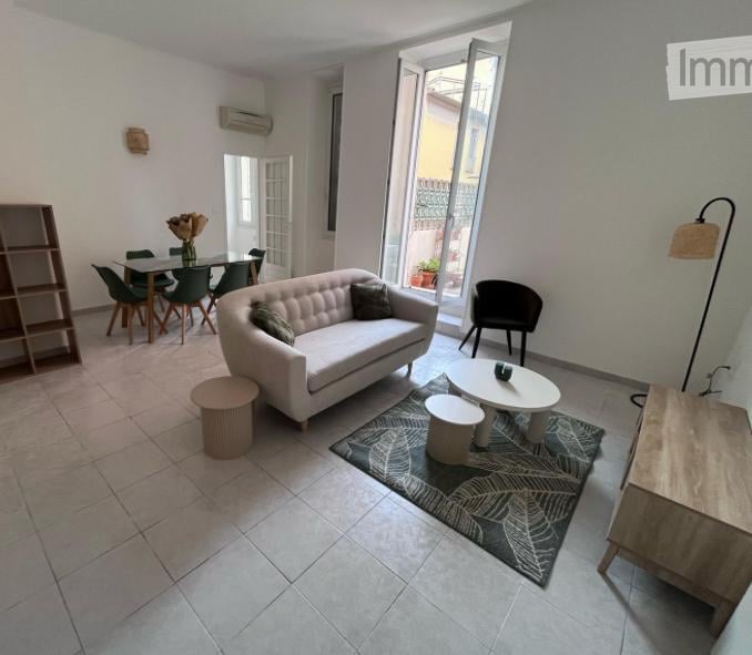 Bright living room with a white sofa, coffee table, dining area, and large windows opening to a balcony. Includes a floor lamp, side tables, and a shelf.