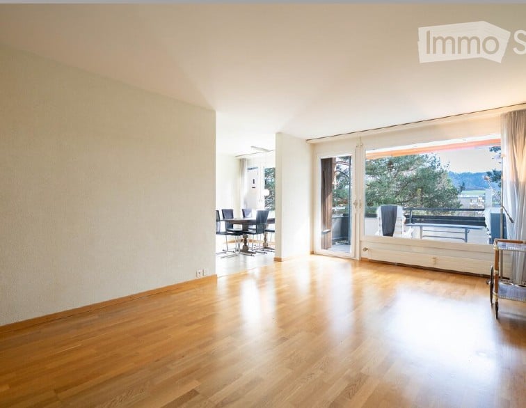 Bright, open living space with wooden flooring, large sliding glass doors leading to a balcony, and a small dining area with a table and chairs.