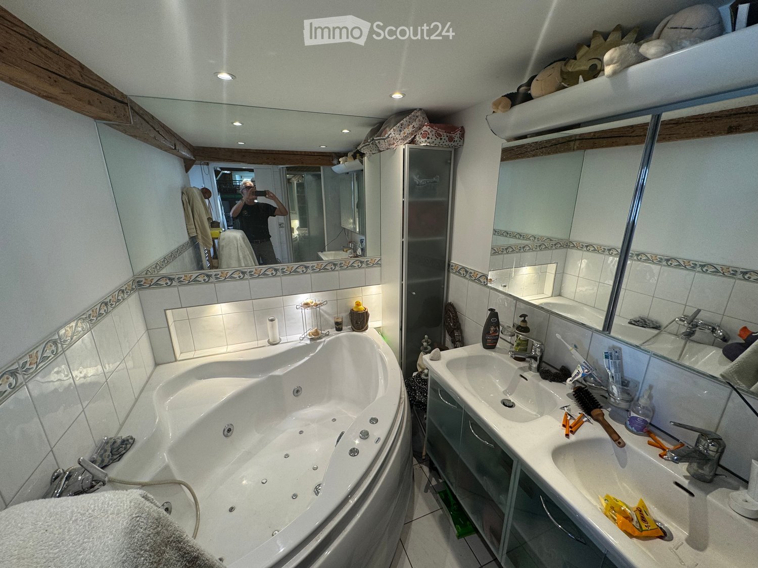 Bathroom with a large bathtub, double sink, mirrors, and various toiletries. The room features tiled walls and a wooden ceiling.