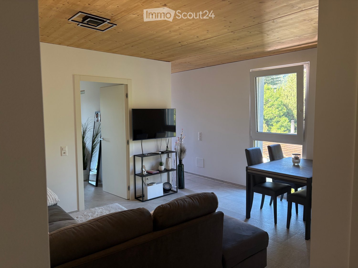 Modern living room with a dark brown sofa, a black dining table with chairs, a shelf with decorative items, and a television mounted on the wall. The room features light-colored walls, a wooden ceiling, and large windows with a view of greenery.