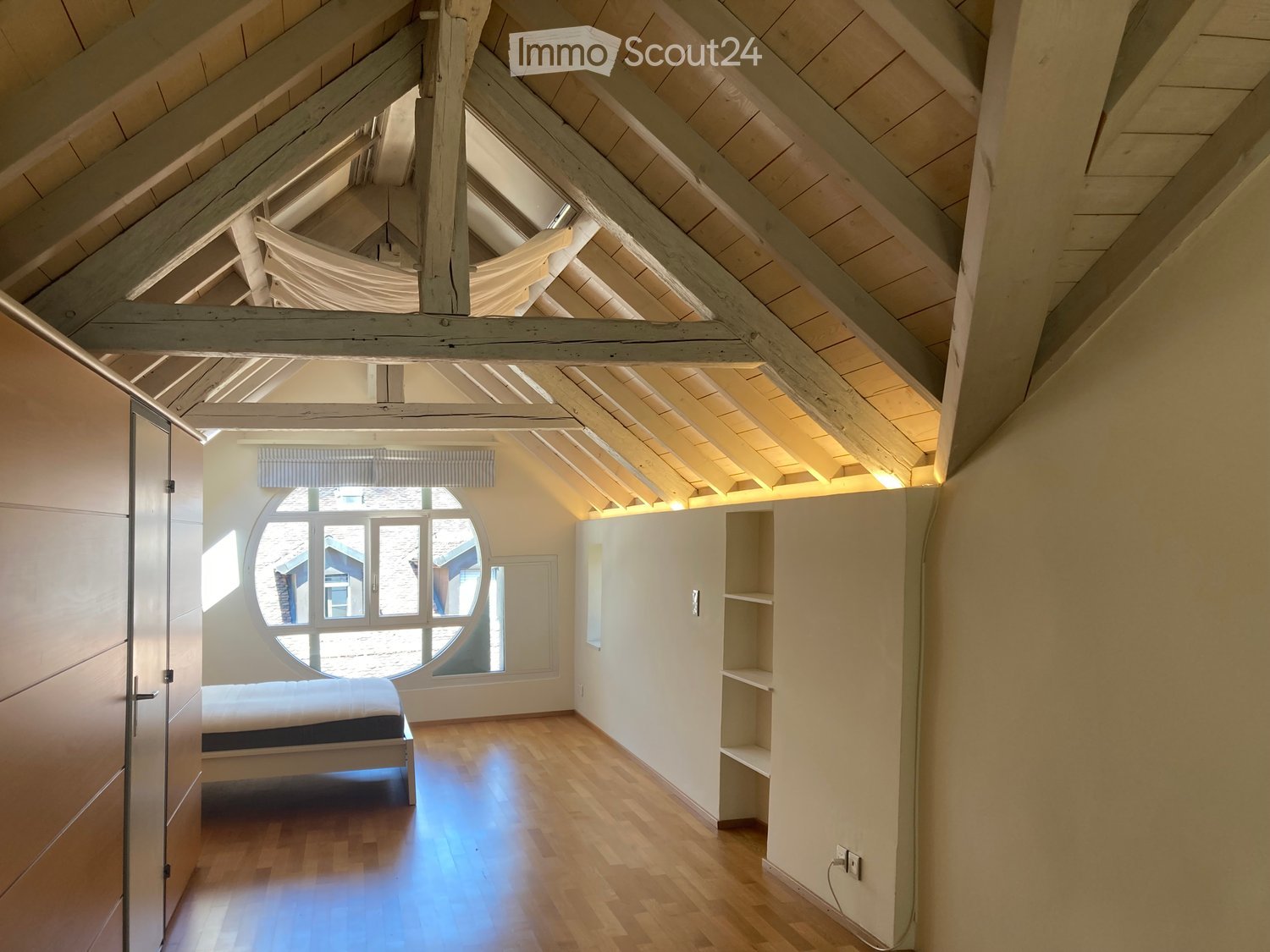 Bright attic bedroom with wooden floors, exposed wooden beams, large round window, built-in shelves, and a simple bed.