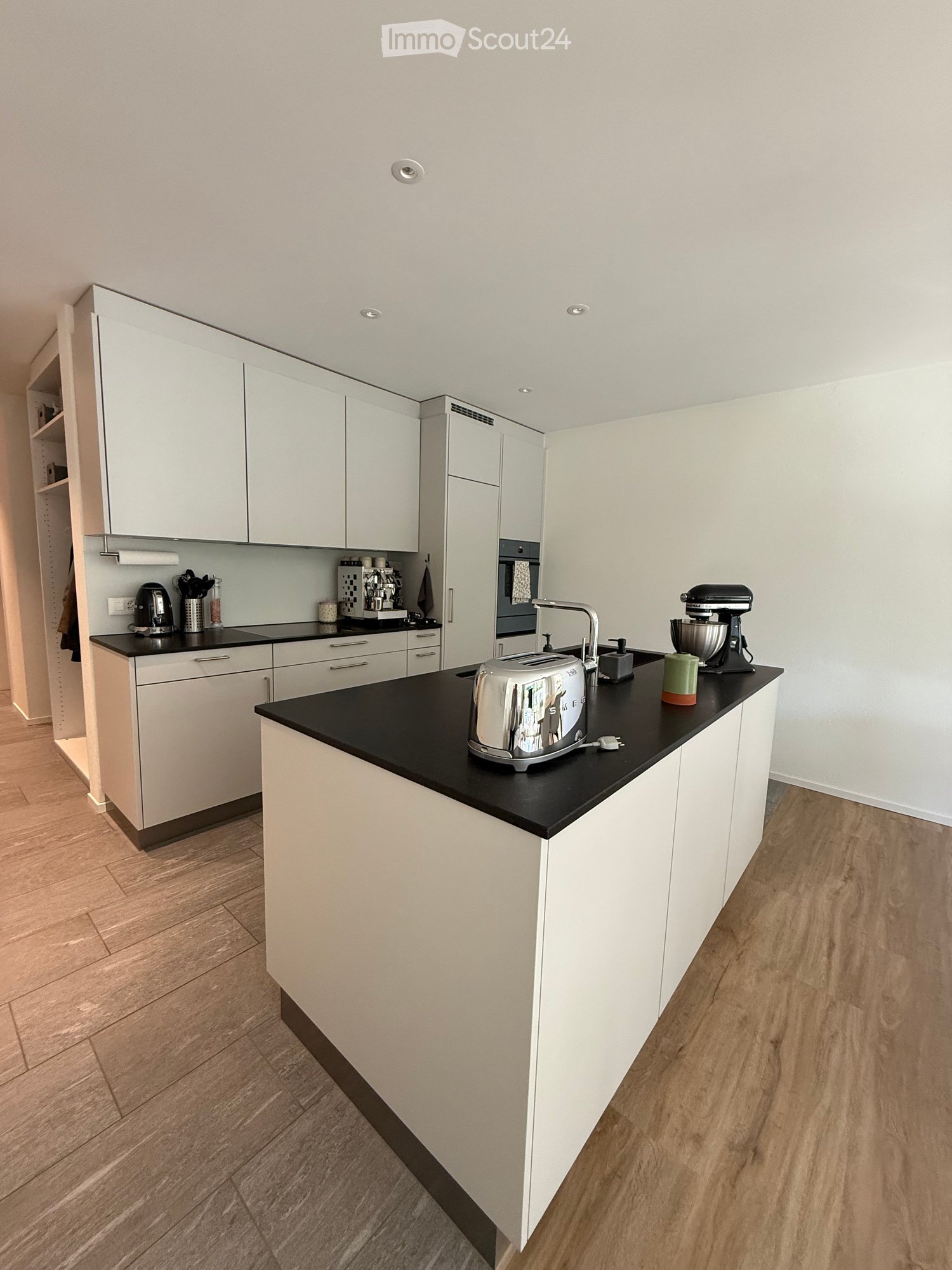 Modern kitchen with white cabinets, black countertops, and wooden flooring. Visible appliances include a toaster, mixer, coffee maker, and oven.