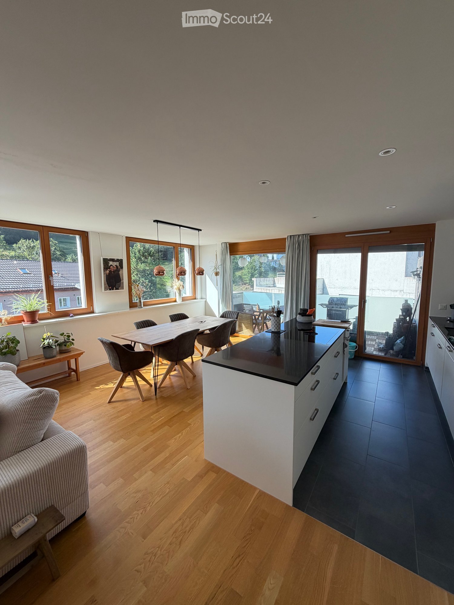 Open-plan living and dining area with wooden flooring, a sofa, dining table with chairs, kitchen island, pendant lights, and large windows with a view of the garden.