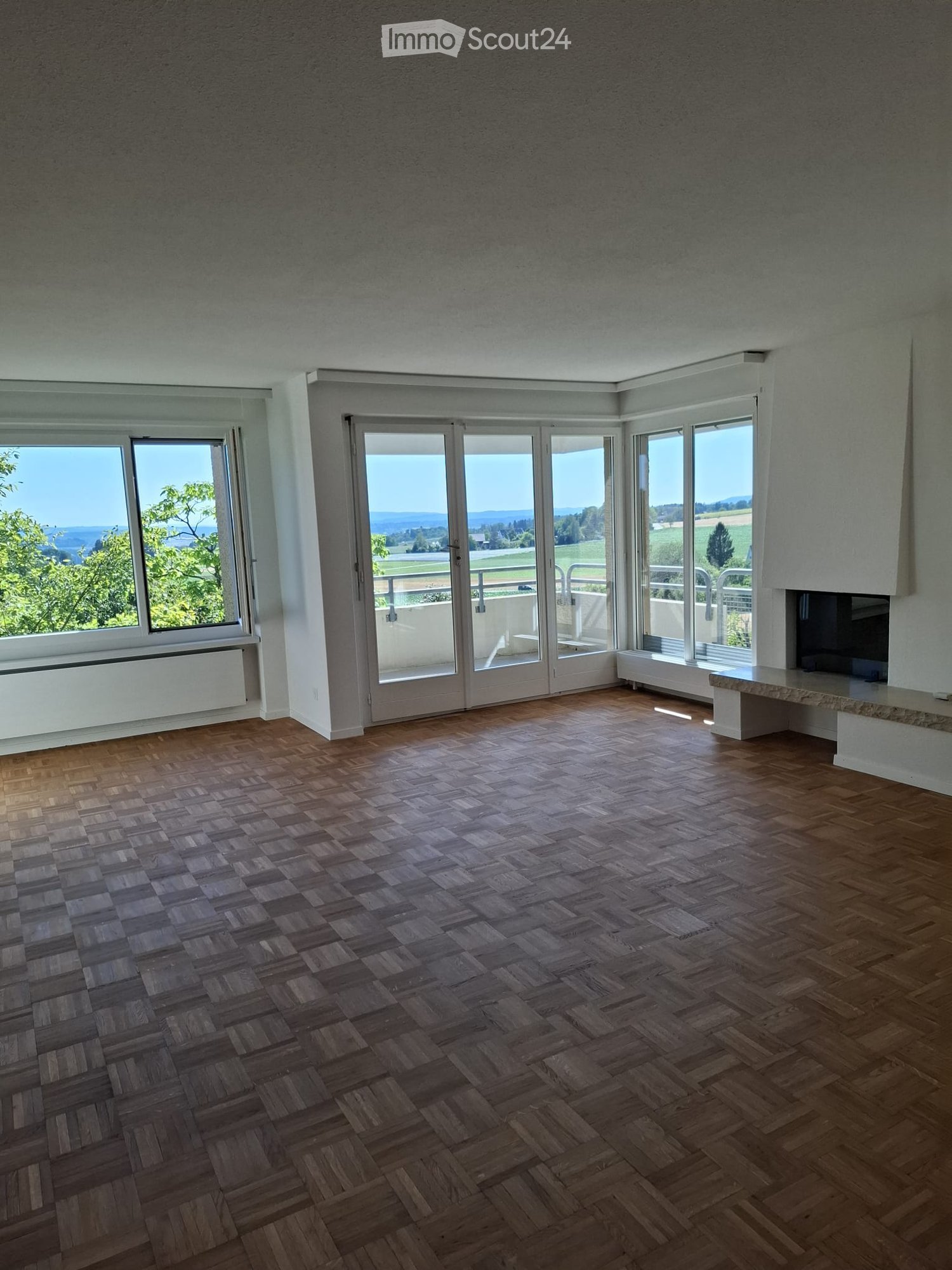 Empty room with wooden herringbone flooring, large windows and sliding glass doors leading to a balcony, offering scenic views of greenery and fields.