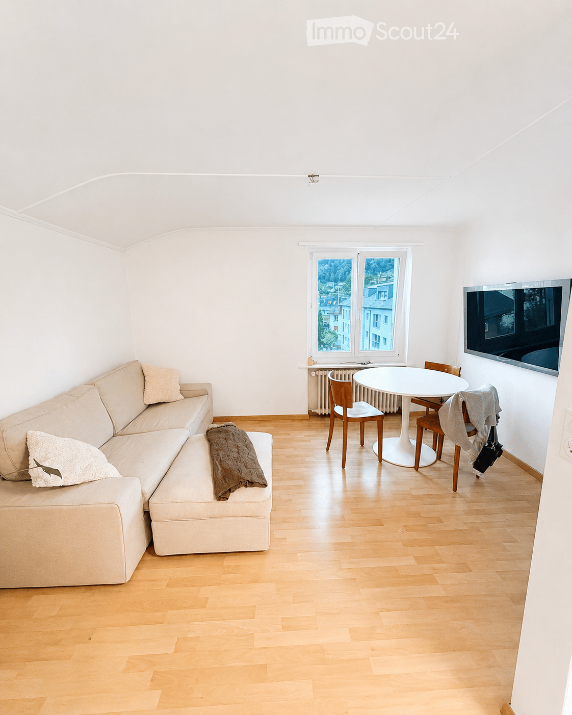 Bright living room with light wooden flooring, a cream-colored sectional sofa, a round white dining table with four chairs, and a flat-screen TV mounted on the wall. The room features large windows with a scenic view.