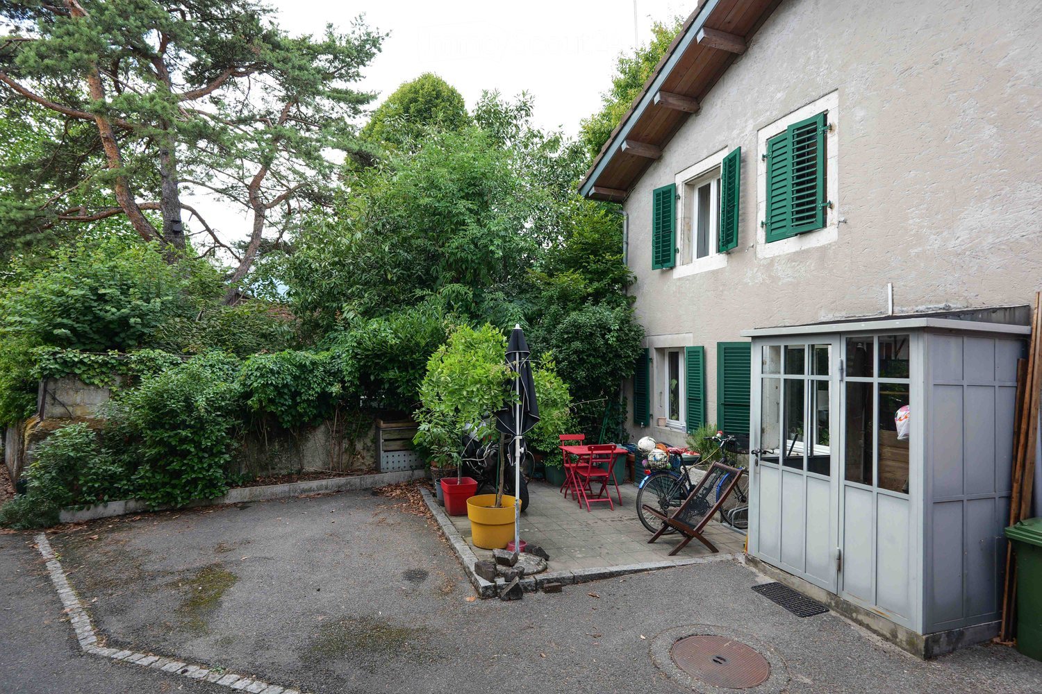 Outdoor area with a small patio, red chairs, a table, bicycles, potted plants, and a shed attached to a house with green shutters.