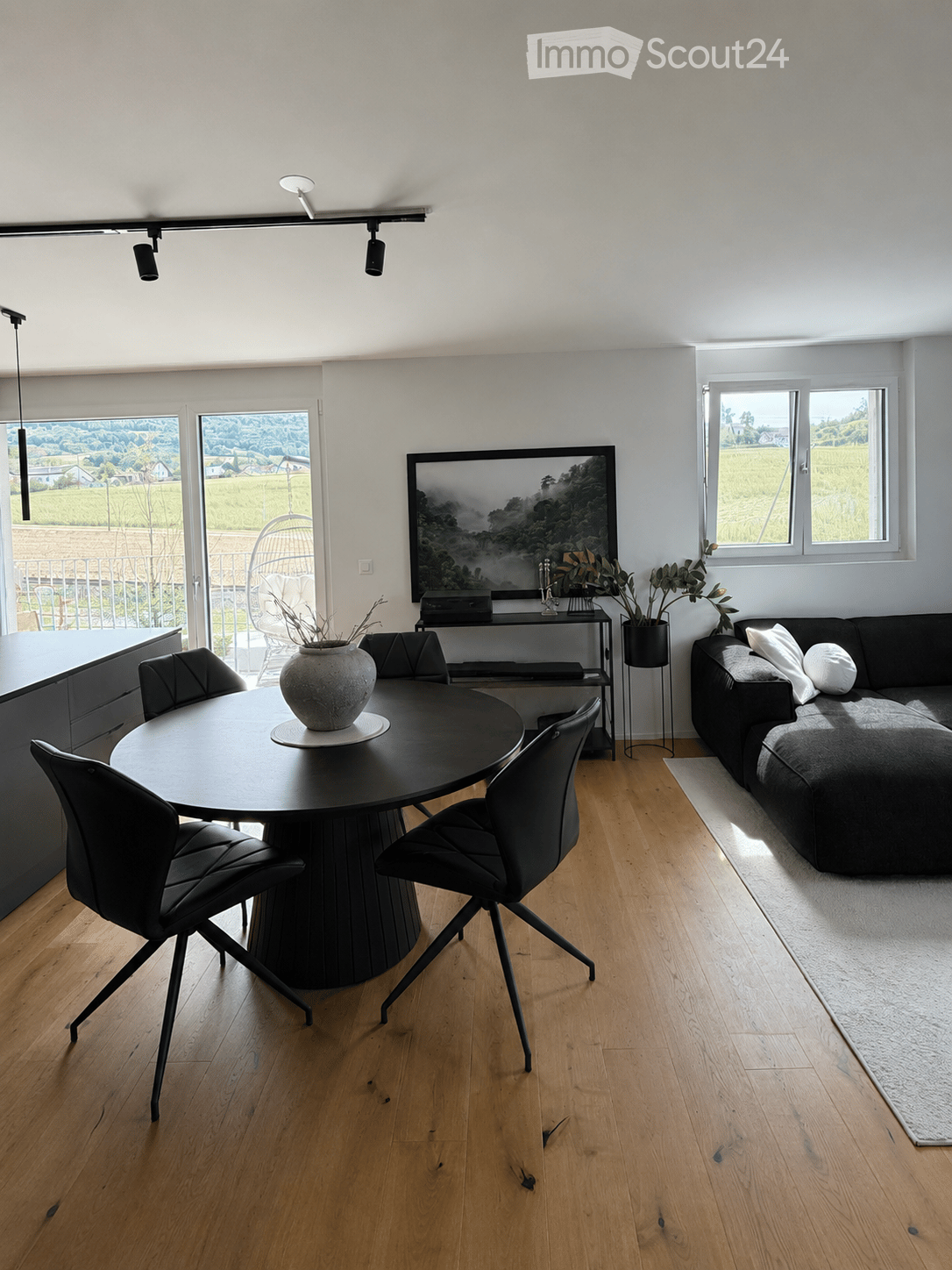 Modern living room with light wooden flooring, a black sofa, a round black coffee table with a vase, and a black dining table with chairs. Large windows offer a scenic view.