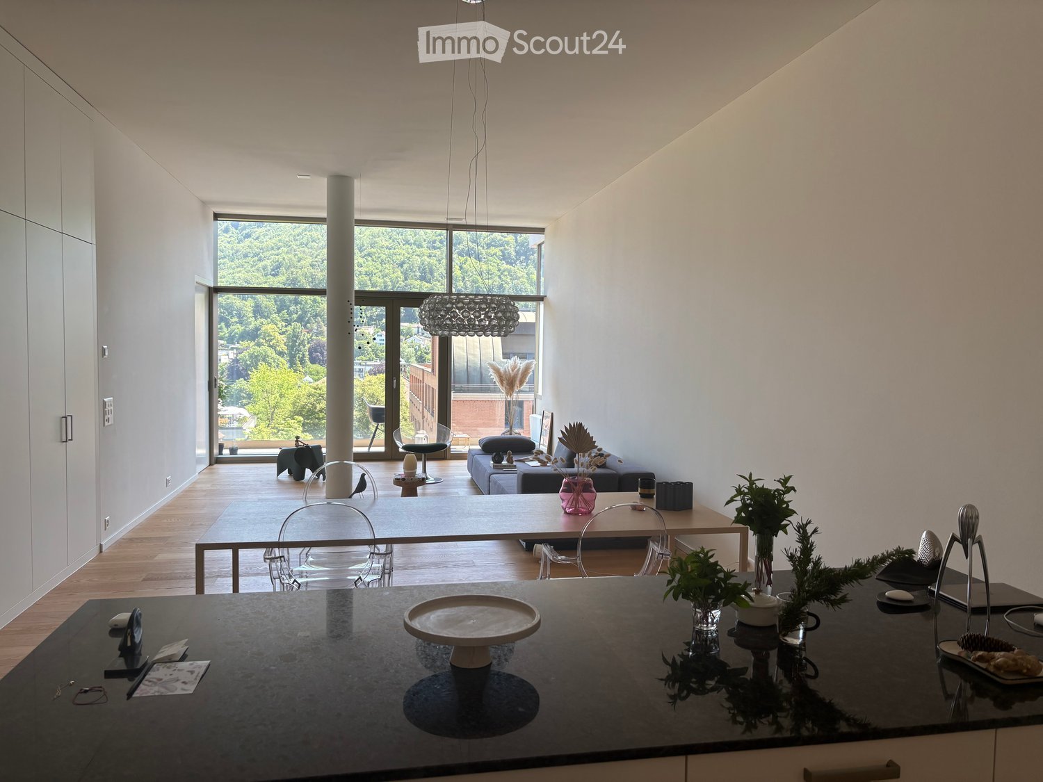 Open-plan living and dining area with modern furniture, large windows, and a chandelier. Includes a dining table, chairs, sofa, and decorative items.