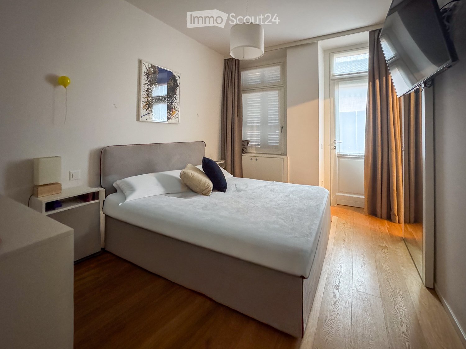 Cozy bedroom with a large bed, nightstand, lamp, and artwork on the wall. Wooden flooring and large windows with curtains.