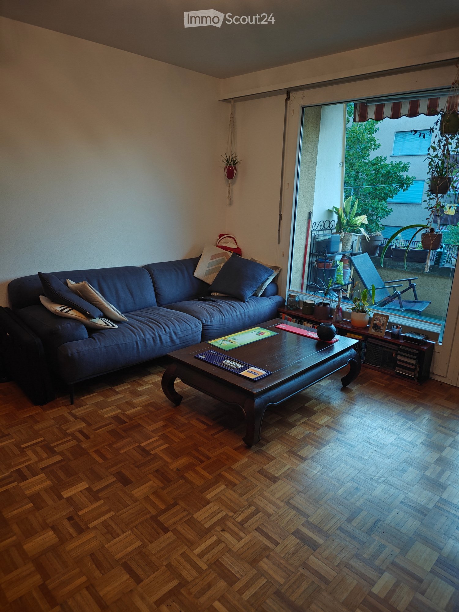 Living room with wooden herringbone flooring, a dark blue sectional sofa, a dark wooden coffee table, and a small table with plants near the balcony entrance.