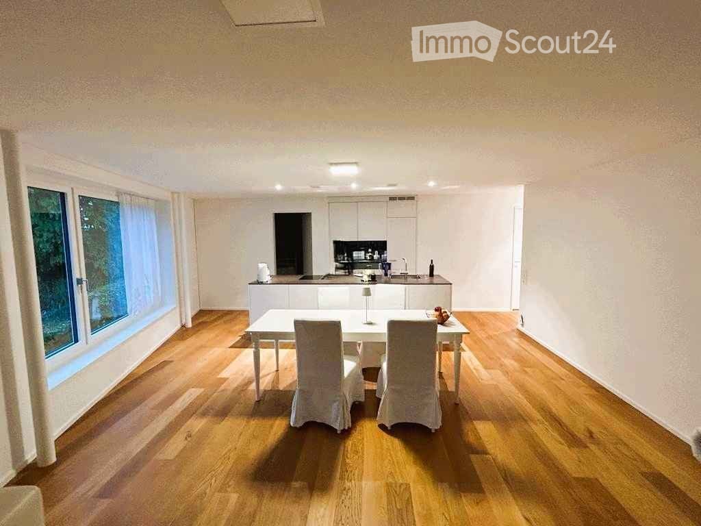 Bright, open-plan kitchen and dining area with wooden flooring, large windows, white cabinetry, and a central dining table with chairs.