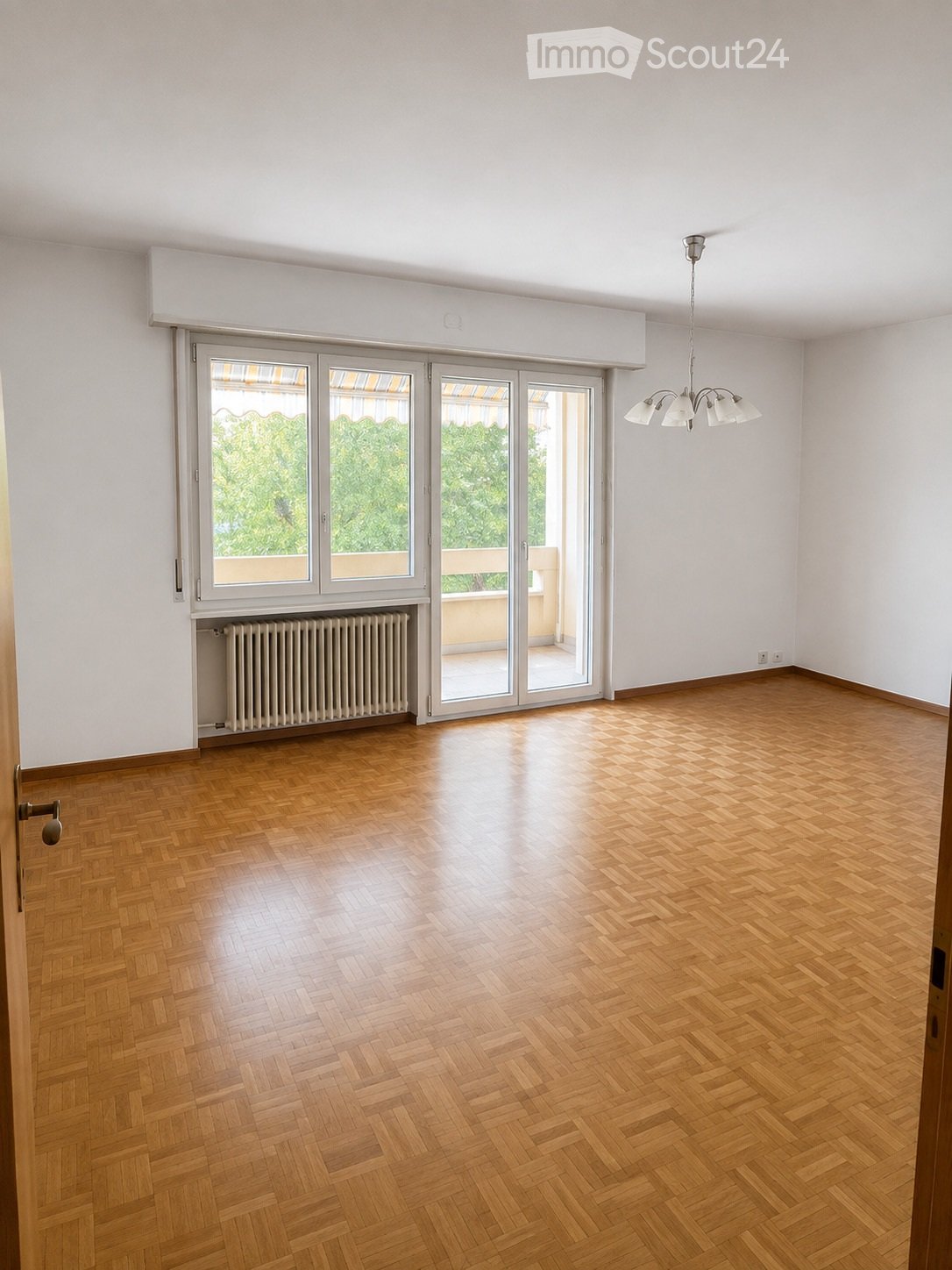 Empty room with wooden herringbone flooring, white walls, large sliding glass doors leading to a balcony, radiator, and a ceiling light fixture.