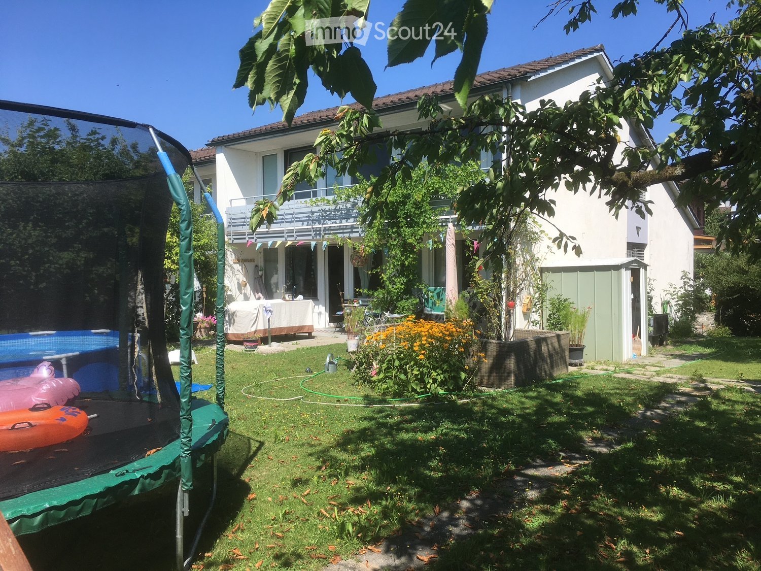 Lush garden with a trampoline, swimming pool, covered patio, and colorful flowers, surrounded by a two-story house with a balcony.