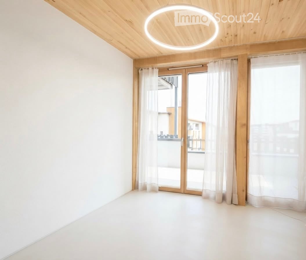 Bright, unfurnished room with white walls, wooden ceiling, large sliding glass doors leading to a balcony, and sheer white curtains.