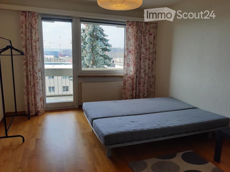 A small, furnished bedroom with a twin-sized bed, wooden flooring, large windows with curtains, and a radiator beneath the windows. A wardrobe is visible on the left side, and a patterned rug is placed near the bed.