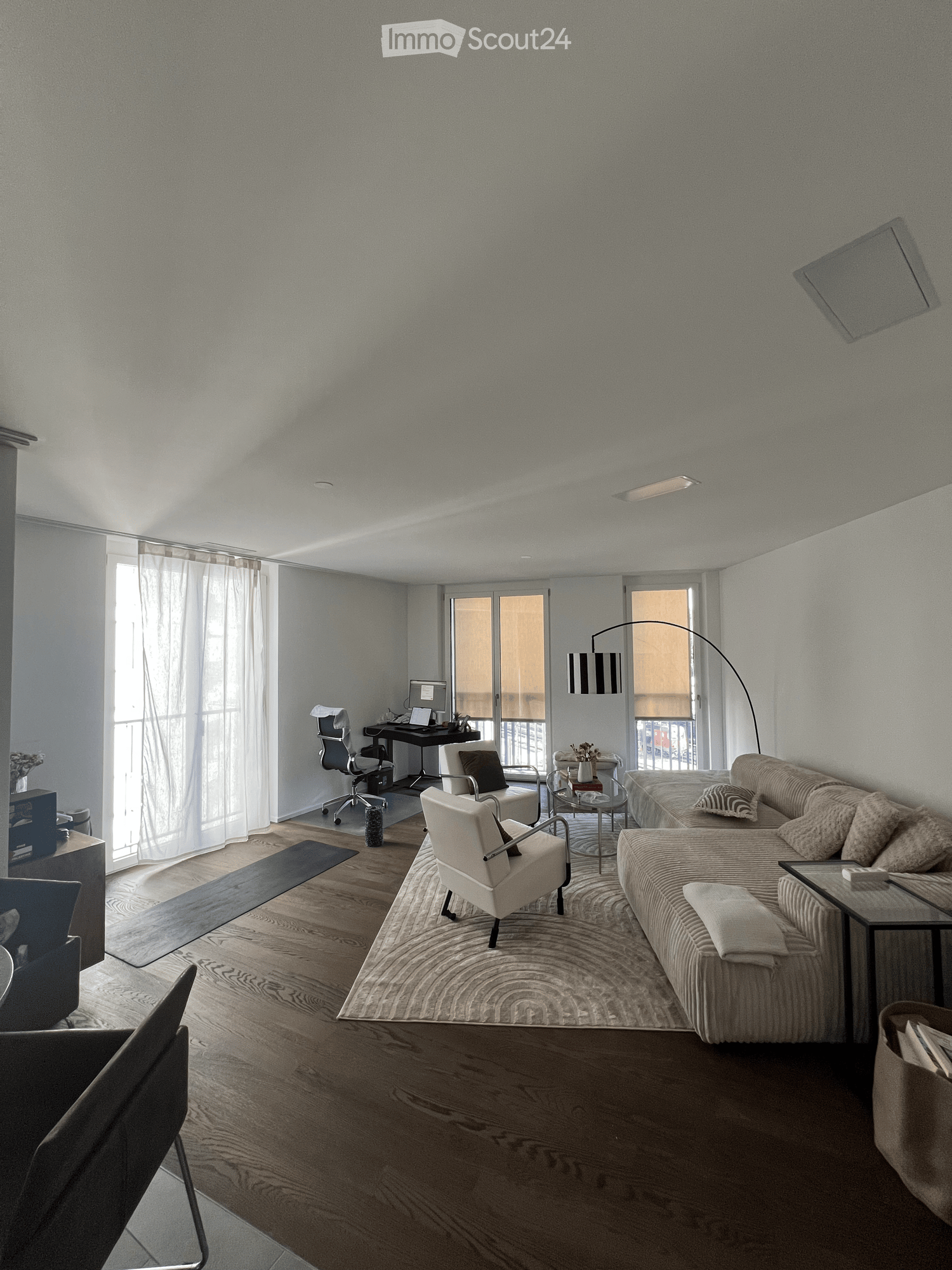 Modern living room with a sofa, chairs, coffee table, side table, floor lamp, and a laptop on a desk near large windows with sheer curtains.