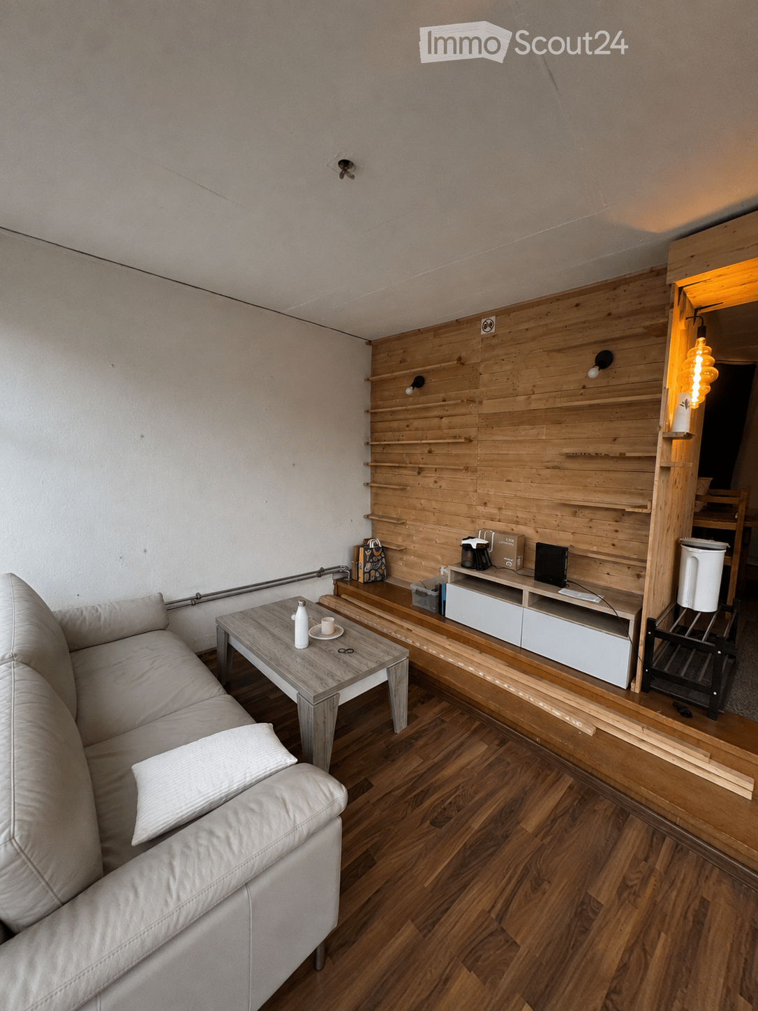 Modern living room with light wooden flooring, white walls, and a wooden accent wall. Features a white leather sofa, a wooden coffee table with a bottle and cup, a TV stand with a laptop, and built-in wooden shelves.
