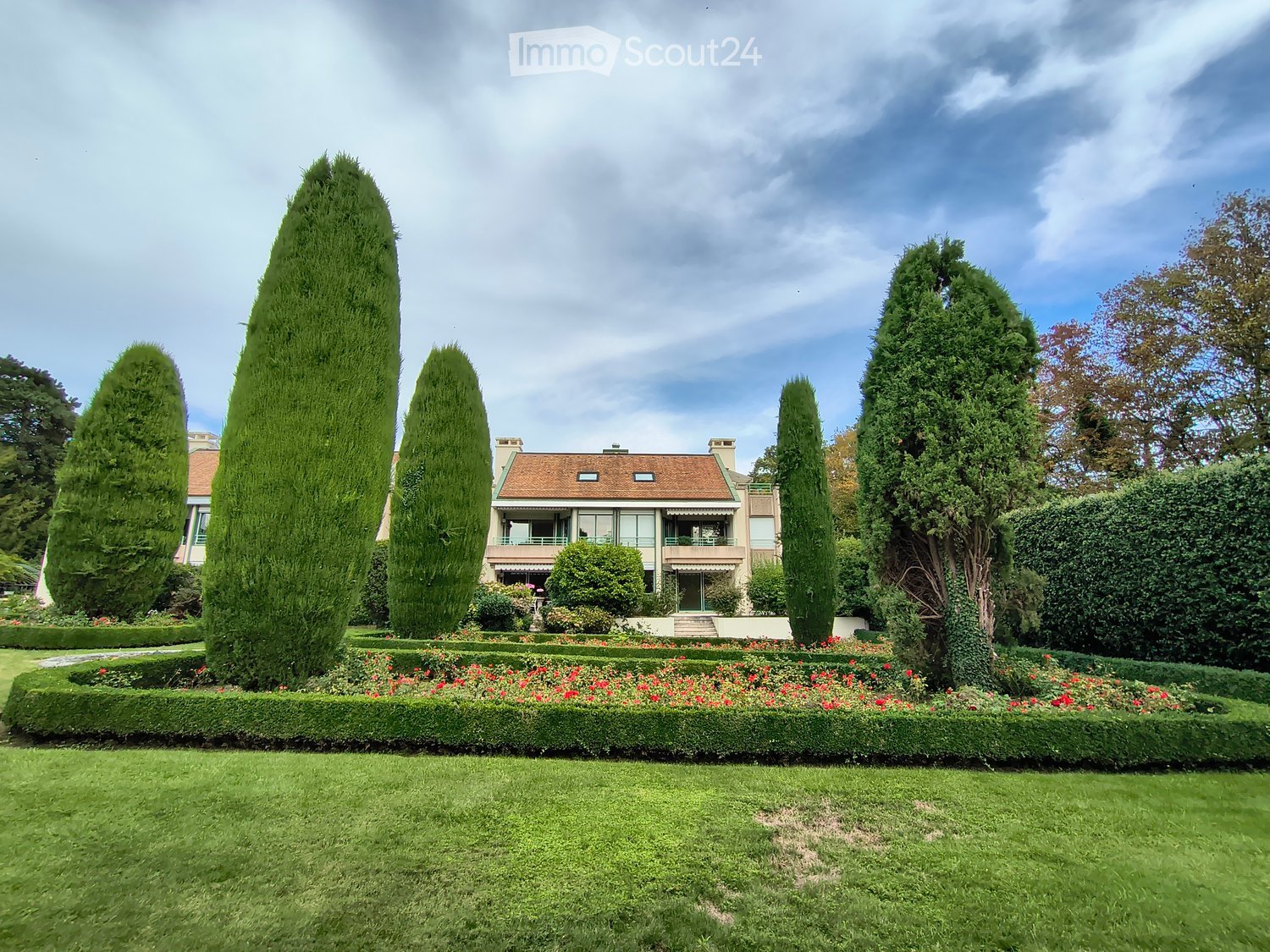 A well-maintained garden with a large house in the background, featuring neatly trimmed hedges, tall coniferous trees, and a variety of flowering plants.