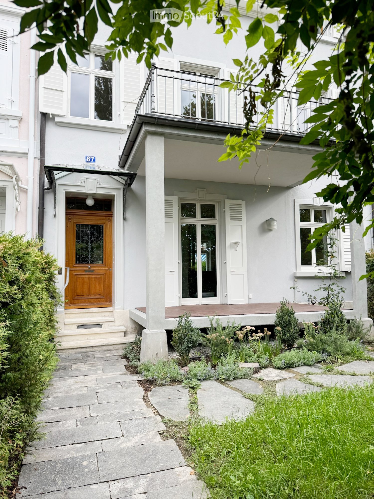 Two-story white building with a wooden front door, balcony, and small garden area with plants and shrubs.